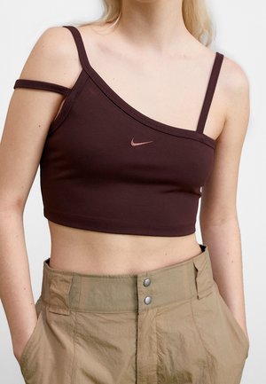Vrouw draagt een asymmetrische crop top in donkerbruin met dunne bandjes en beige hoge taillebroek, handen in de zakken.