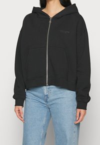 Svart croppad hoodie i bomull, med dragkedja fram, kängurufickor och fodrad huva. Mjuk textur med en minimalistisk design.