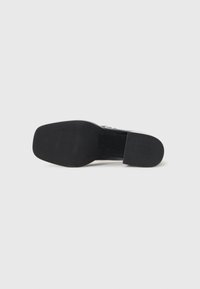 Semelle de chaussure noire avec un bout carré et un petit talon reposant sur une surface blanche uniforme, montrant le motif de la semelle.