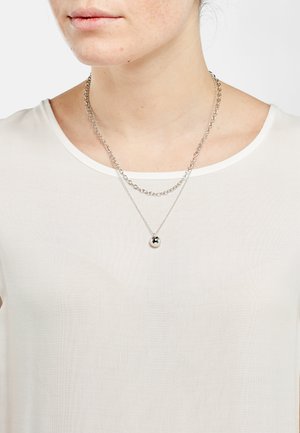 Collier en argent composé de deux chaînes superposées ; l'une d'elles est ornée d'un pendentif sphérique lisse. Porté sur un top blanc léger et semi-transparent.