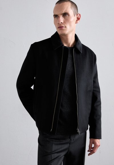 Black wool-style jacket with a pointed collar, front zip closure, and side pockets, worn over a dark sweater and tailored pants.