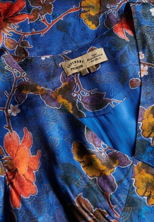 Blå mønstret stof med blomsterdesign i orange, gul og lilla. Inkluderer et synligt mærke med "SUPERDRY" og "PREMIUM" branding.