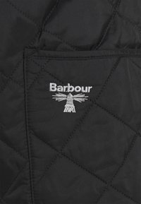 Tissu noir matelassé avec motif en losanges, présentant un logo Barbour tissé en argent. Détail d'une poche visible sur le côté droit.