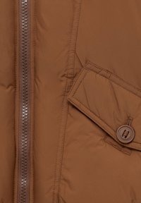 Gros plan d'une veste en tissu marron avec une fermeture éclair à gauche et une poche à rabat boutonnée à droite.