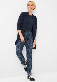 Marine cardigan over een donkerblauw top, gecombineerd met geblokte broek in blauw en zwart. Schoeisel heeft zwarte accenten en witte veters.
