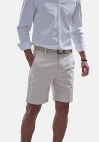 Hellbeige Baumwollshorts mit zwei seitlichen Taschen und einem Reißverschluss. Getragen mit einem weißen Langarmhemd und einem geflochtenen Gürtel.