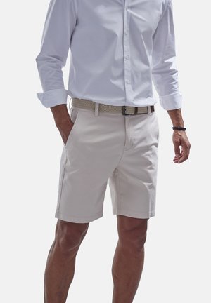 Light beige shorts made of cotton with two side pockets and a zipper fly. Worn with a white long-sleeve shirt and a woven belt.