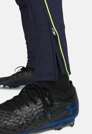 Marineblauwe sportbroek met een zijrits en groene bies, gecombineerd met zwarte, getextureerde voetbalschoenen met blauwe accenten.