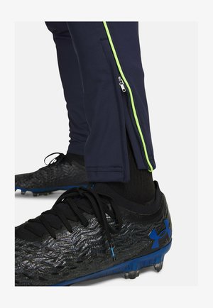 Marineblauwe sportbroek met een zijrits en groene bies, gecombineerd met zwarte, getextureerde voetbalschoenen met blauwe accenten.