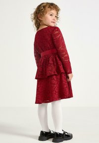 Rød blondekjole med lange ærmer, en peplum kant og et matchende bælte. Hvide strømpebukser og sorte patenterede sko fuldender outfittet.