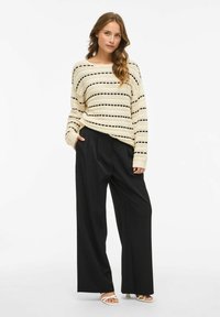 Off-white pleten pulover s črnimi črtami v kombinaciji z črnimi širokimi hlačami z visokim pasom. Model prikazuje sproščen kroj in sproščen stil.
