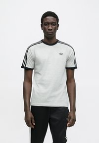 Γκρι αθλητική μπλούζα με μαύρες λεπτομέρειες, κοντά μανίκια και στρογγυλή λαιμόκοψη. Διαθέτει τρεις μαύρες ρίγες σε κάθε ώμο και ένα μικρό λογότυπο.