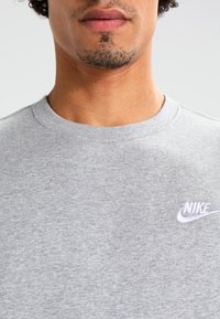 Harmaa puuvillasekoitekankainen pyöreäkauluksinen collegepaita, jossa on pehmeä tekstuuri; rinnassa valkoinen kirjailtu Nike-logo.