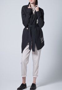 Grafitowy dzianinowy kardigan ze sznurkiem do wiązania, o prążkowanej strukturze i geometrycznych wzorach na krawędzi, zestawiony z jasnobeżowymi spodniami i czarnymi butami.