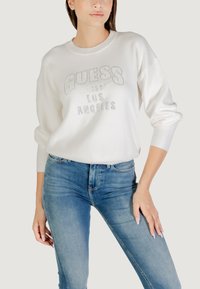 Witte sweatshirt met geperste "GUESS" tekst, ronde halslijn en driekwartmouwen, gecombineerd met blauwe skinny jeans.