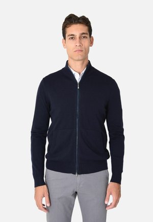 Maglione navy con zip in un morbido tessuto a maglia, colletto alto, polsini a coste e due tasche frontali, indossato sopra una camicia bianca con colletto.