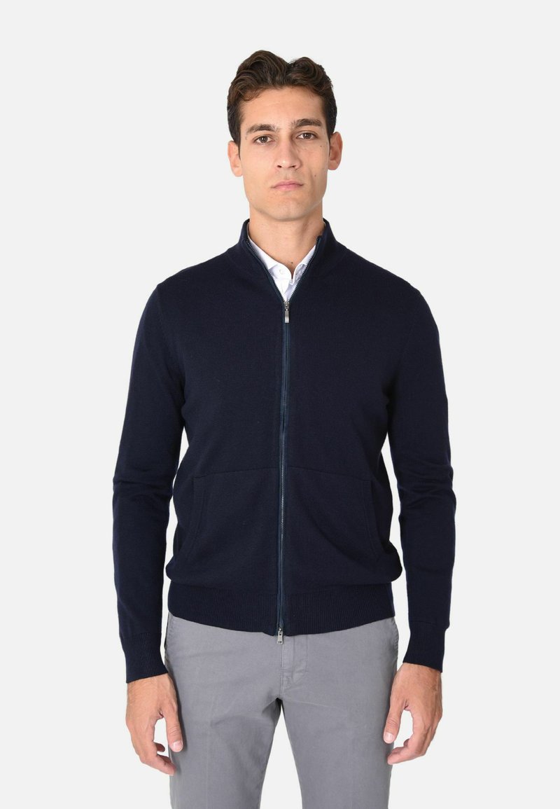 Maglione navy con zip in un morbido tessuto a maglia, colletto alto, polsini a coste e due tasche frontali, indossato sopra una camicia bianca con colletto.