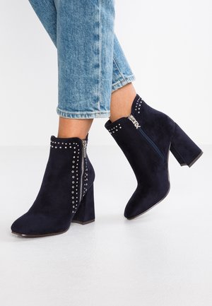 Woman wearing blue suede ankle boots with block heels, silver studs, and side zippers paired with cropped light blue jeans.