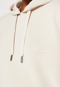 Sudadera beige con una tela suave y texturizada. Presenta un logo con un acabado blanco brillante y cordones con puntas metálicas.