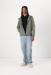 Jachetă ușoară, cu fermoar, de culoare verde măslinie, cu glugă, purtată peste un polo negru, combinată cu blugi albaștri deschiși și pantofi sport bej. Design simplu.