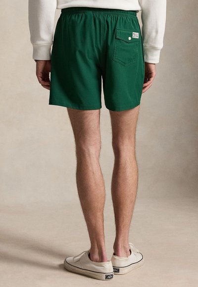 Homme debout, portant un short vert foncé avec une poche arrière et des baskets blanches, visible de la taille aux pieds sur un fond neutre.