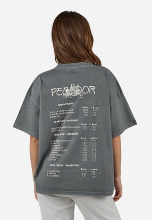 Osoba o brązowych włosach nosząca szarą, za dużą koszulę z listą rodzajów wina i cenami, widziana od tyłu na białym tle.