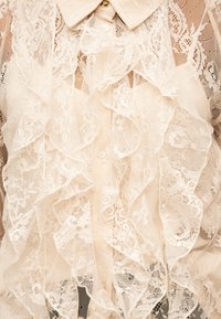 Μπλούζα από μπεζ δαντέλα με λουλουδάτα σχέδια, στρώσεις με βολάν στο μπροστινό μέρος, γιακάς με κουμπιά και διαφανή υφή υφάσματος.