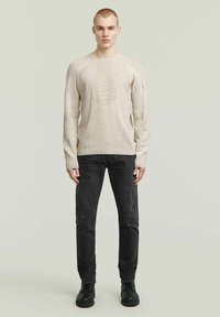 Maglione beige lavorato a maglia con motivi texture e dettagli a costine, abbinato a jeans neri strappati e scarpe nere.