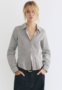 Camicia a righe bianche e nere con colletto rigido, maniche lunghe e forma aderente, abbinata a jeans in denim scuro.