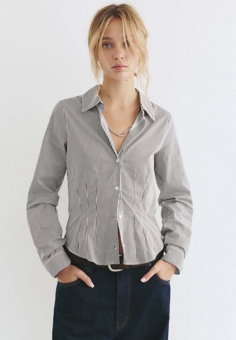 Camicia a righe bianche e nere con colletto rigido, maniche lunghe e forma aderente, abbinata a jeans in denim scuro.