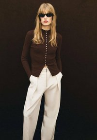 Cárdigan marrón con acanalado y botones en la parte delantera, combinado con pantalones de pierna ancha blancos que presentan un detalle de cremallera. Unas gafas de sol negras completan el look.