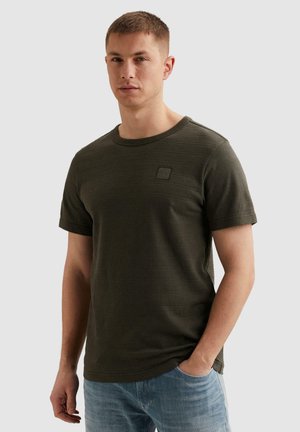 Man met kort haar, gekleed in een donker groen, gestructureerd kort shirt en lichtblauwe spijkerbroek, staand met één hand in de zak.