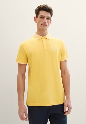 Camisa polo amarilla con cuello clásico, mangas cortas y un sutil logotipo en el pecho. Tela suave con un acabado de color sólido.
