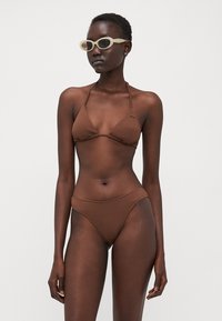 Brun, texturerad bikini med triangulärt behå och lågt skurna byxor. Modell som bär ljusfärgade solglasögon, stående mot en enfärgad bakgrund.