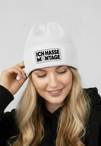 White knit beanie featuring a black rectangular patch with the text "ICH HASSE MONTAGE" and a cat graphic, set against a light background.