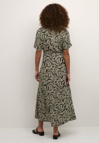 Schwarzes Kleid mit weißem Blattmuster, kurzen Ärmeln und einer fließenden Silhouette. Strukturierten Stoff. Getragen mit schwarzen Blockabsatzsandalen.
