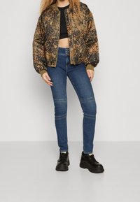 Chaqueta bomber de estampado de leopardo en marrón y negro, combinada con jeans ajustados azules con detalles en las rodillas, y zapatos negros con suela gruesa.