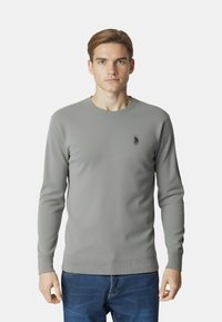 Maglione grigio a maniche lunghe realizzato in tessuto liscio, con collo rotondo a coste e polsini, con un piccolo logo sul petto sinistro. Jeans visibili.