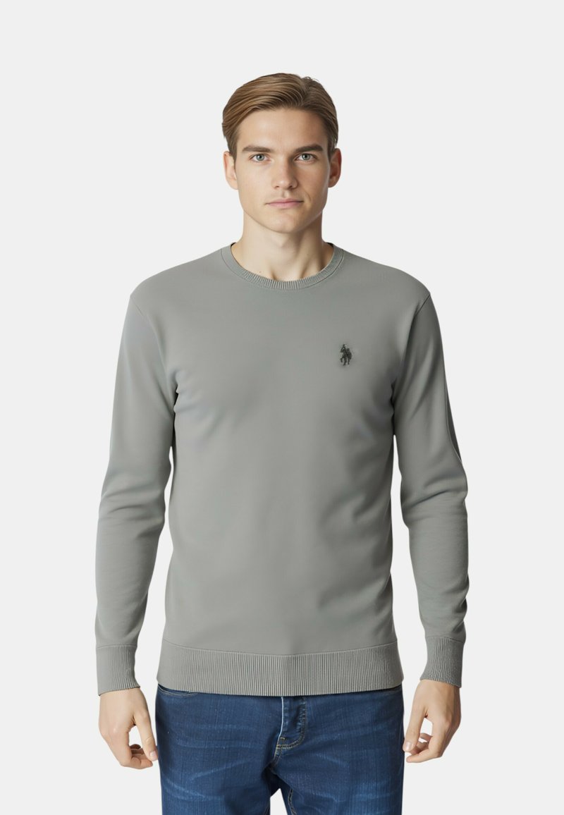 Maglione grigio a maniche lunghe realizzato in tessuto liscio, con collo rotondo a coste e polsini, con un piccolo logo sul petto sinistro. Jeans visibili.