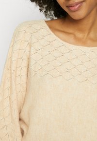 Femme portant un pull en laine beige avec un motif en losange sur le haut de la poitrine et les manches, souriant doucement.