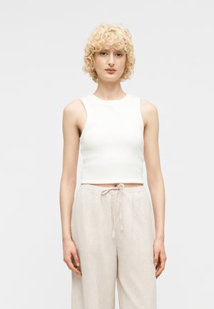 Άτομο με ξανθά, σγουρά μαλλιά που φοράει λευκό αμάνικο crop top και ανοιχτό μπεζ παντελόνι με κορδόνι, στέκεται μπροστά σε απλό φόντο.