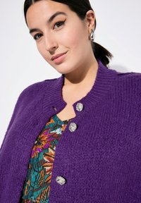 Lila gestrickte Strickjacke aus strukturiertem Stoff, mit silbernen Blumenknöpfen. Über einem bunten, floral gemusterten Oberteil getragen.