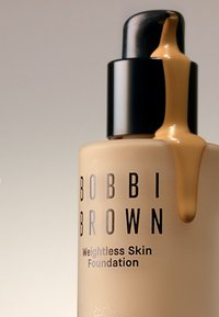 Bobbi Brown Weightless Skin Foundation i en frostet glassflaske med svart pumpe, flytende foundation som renner nedover siden, lys beige farge.