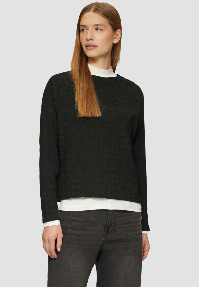Zwarte textuur lange mouwen top met een boxy fit, gelaagd over een wit shirt, aangevuld met grijze jeans. Eenvoudig, hedendaags ontwerp.