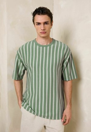 Junger Mann mit grün-weiß gestreiftem, kurzärmeligem Hemd und hellbeiger Hose, der vor einer hellen Wand steht.