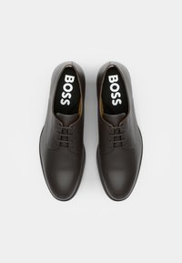 Une paire de chaussures de ville en cuir noir à lacets avec "BOSS" imprimé en blanc sur les semelles intérieures noires, présentée de dessus sur un fond blanc.