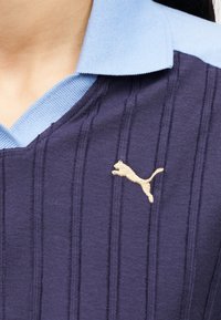 Κοντινό πλάνο ενός ατόμου που φοράει μπλε σκούρο ριπ μπλουζάκι με ανοιχτό μπλε γιακά και μικρό χρυσό λογότυπο Puma στο στήθος.