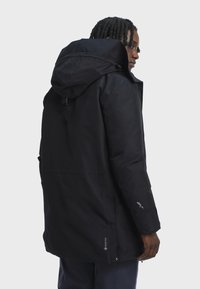 Person with dark braided hair wearing a black Gore-Tex hooded jacket seen from the back and side on a light gray background.