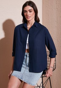 Camicia blu navy con bottoni e maniche arrotolate, realizzata in tessuto morbido, indossata sopra un crop top bianco e abbinata a una gonna mini in denim.