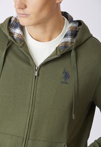 Felpa con zip di colore verde oliva, con rivestimento a quadri nel cappuccio. Presenta un logo ricamato, tasche frontali e una texture liscia.